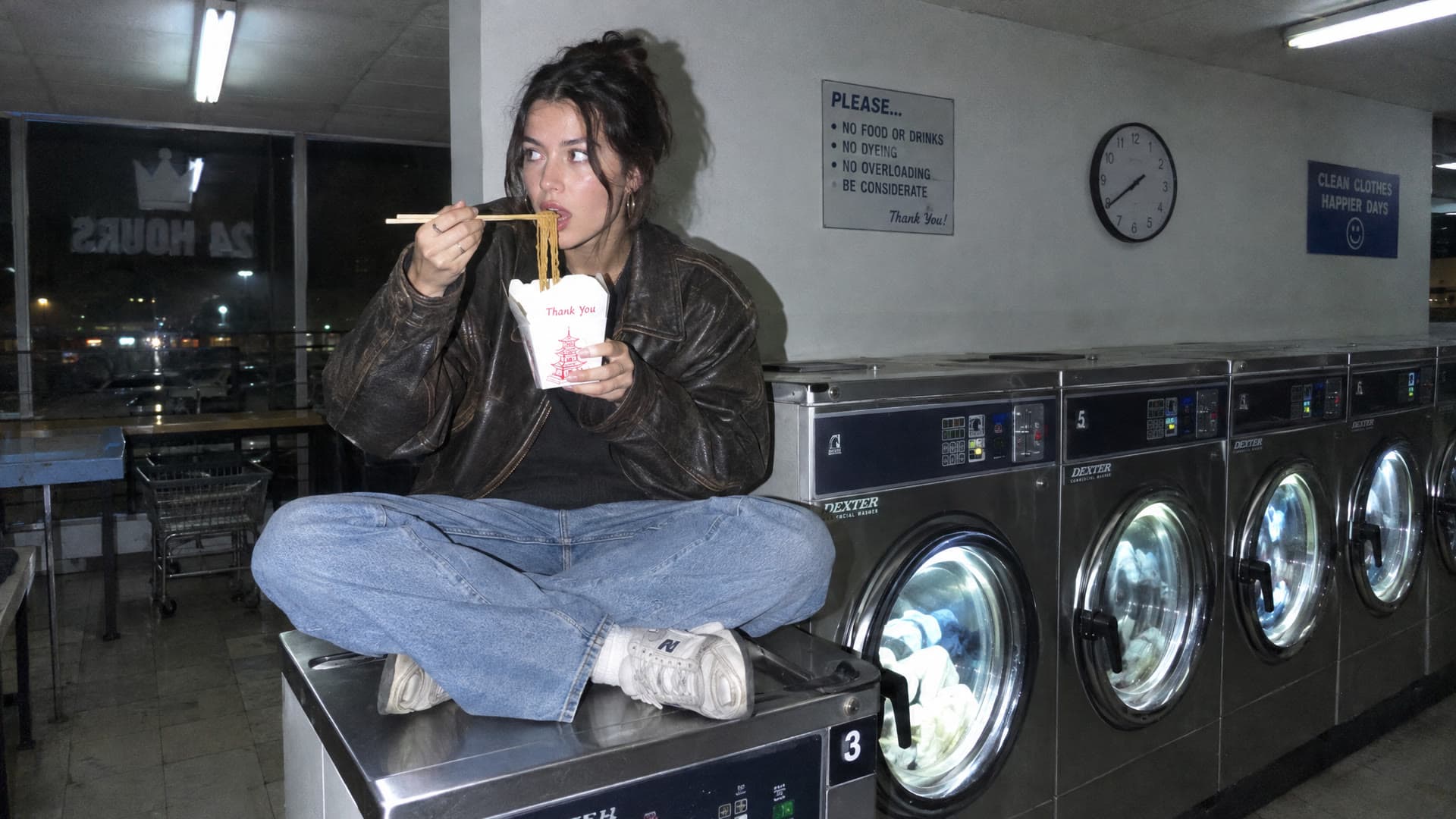 Flash photo of a woman eating noodles on a laundromat washer, the start frame for a Kling 4 clip