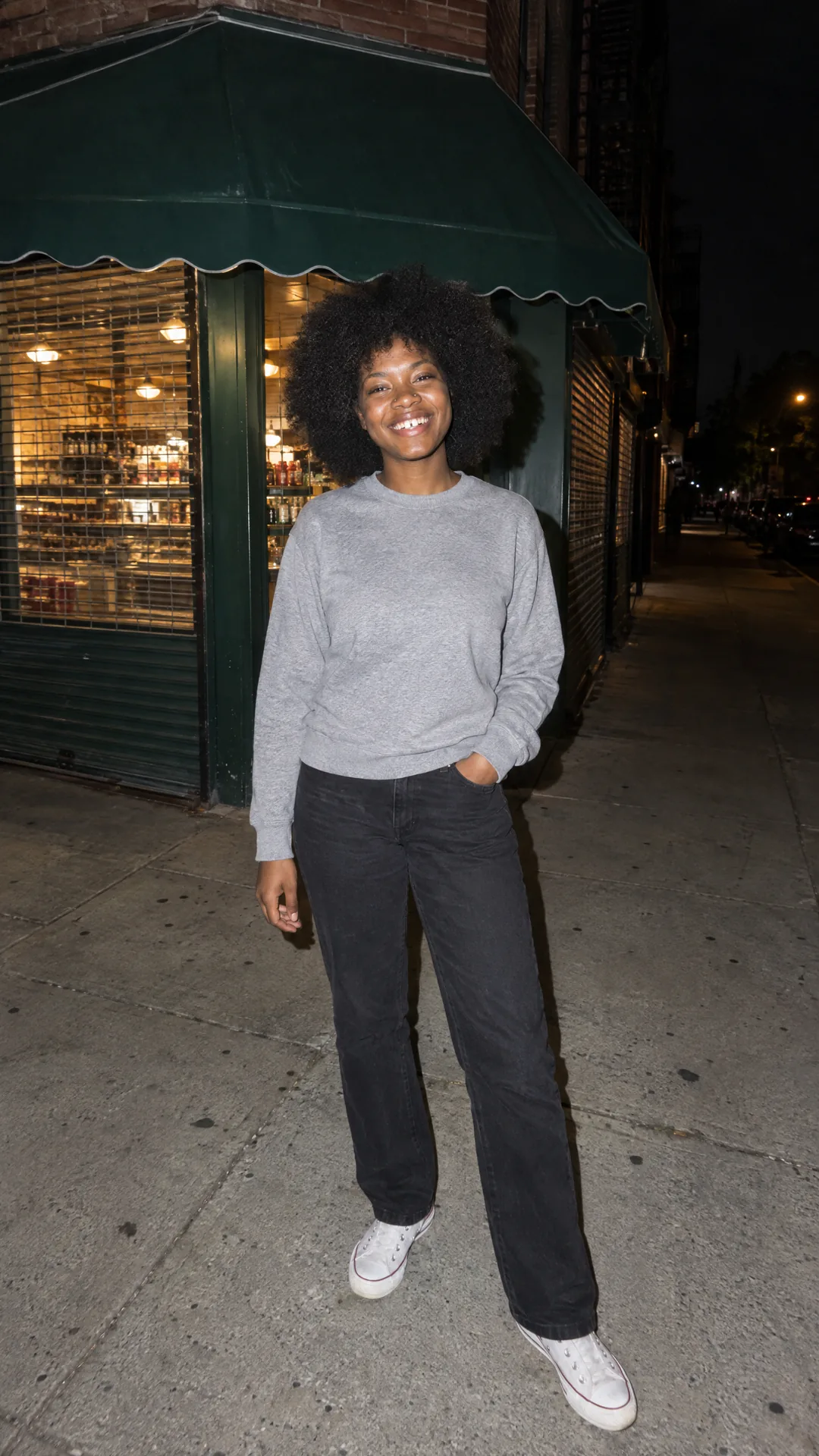 A night flash photo of a woman with a big natural afro and a gap-toothed grin on a city sidewalk, in a grey sweatshirt, black jeans and white sneakers